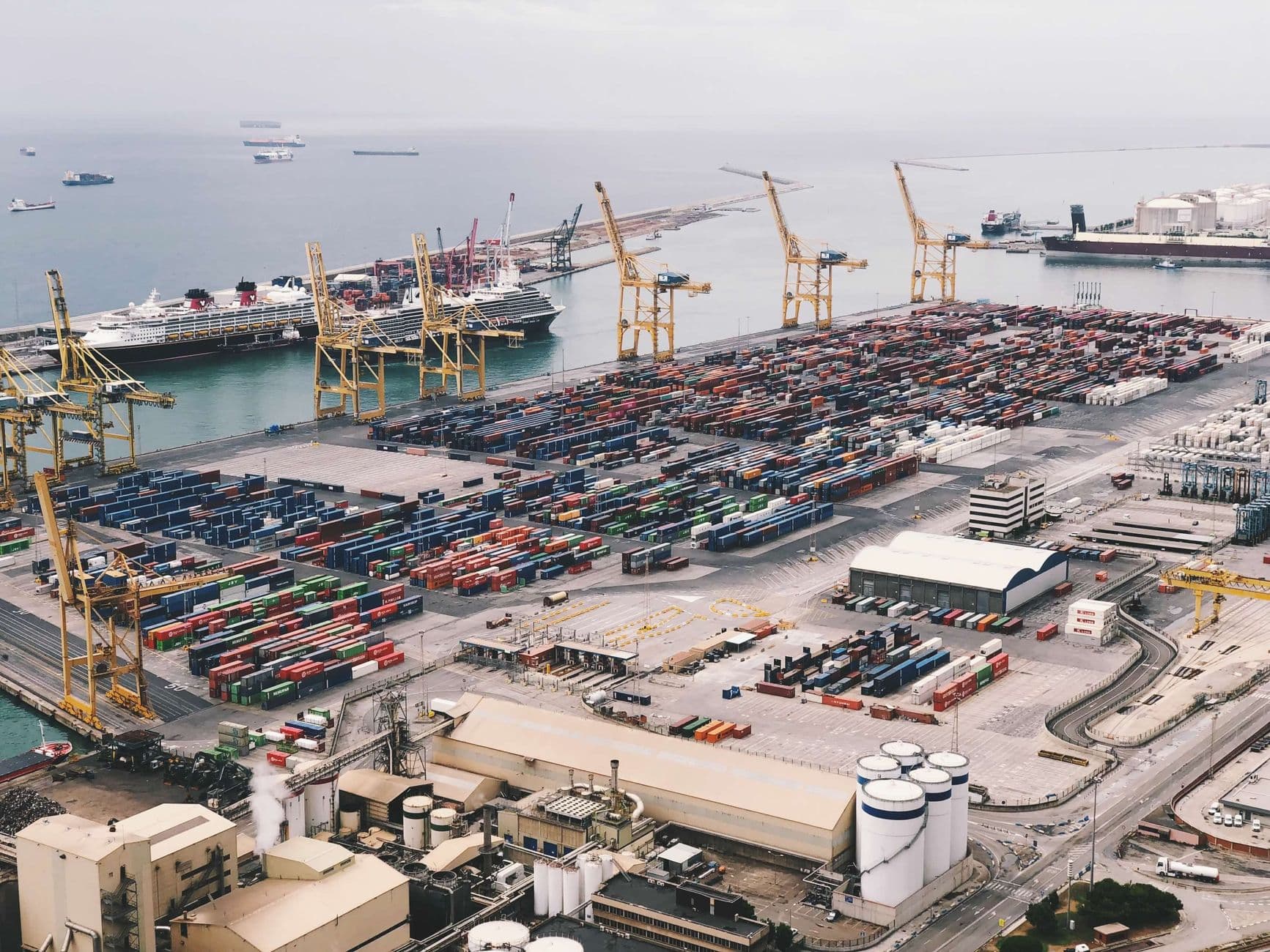 Expansive aerial view of a bustling container port with large cranes and cargo ships under a cloudy sky.